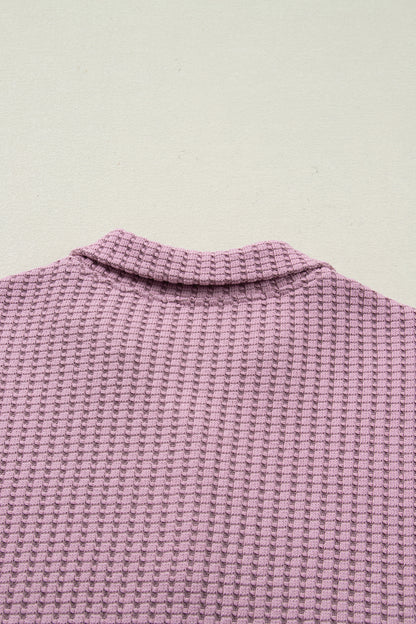 Woman wearing a Blush textured waffle knit pullover with snap collar, relaxed long sleeves, and ribbed cuffs with white pants.